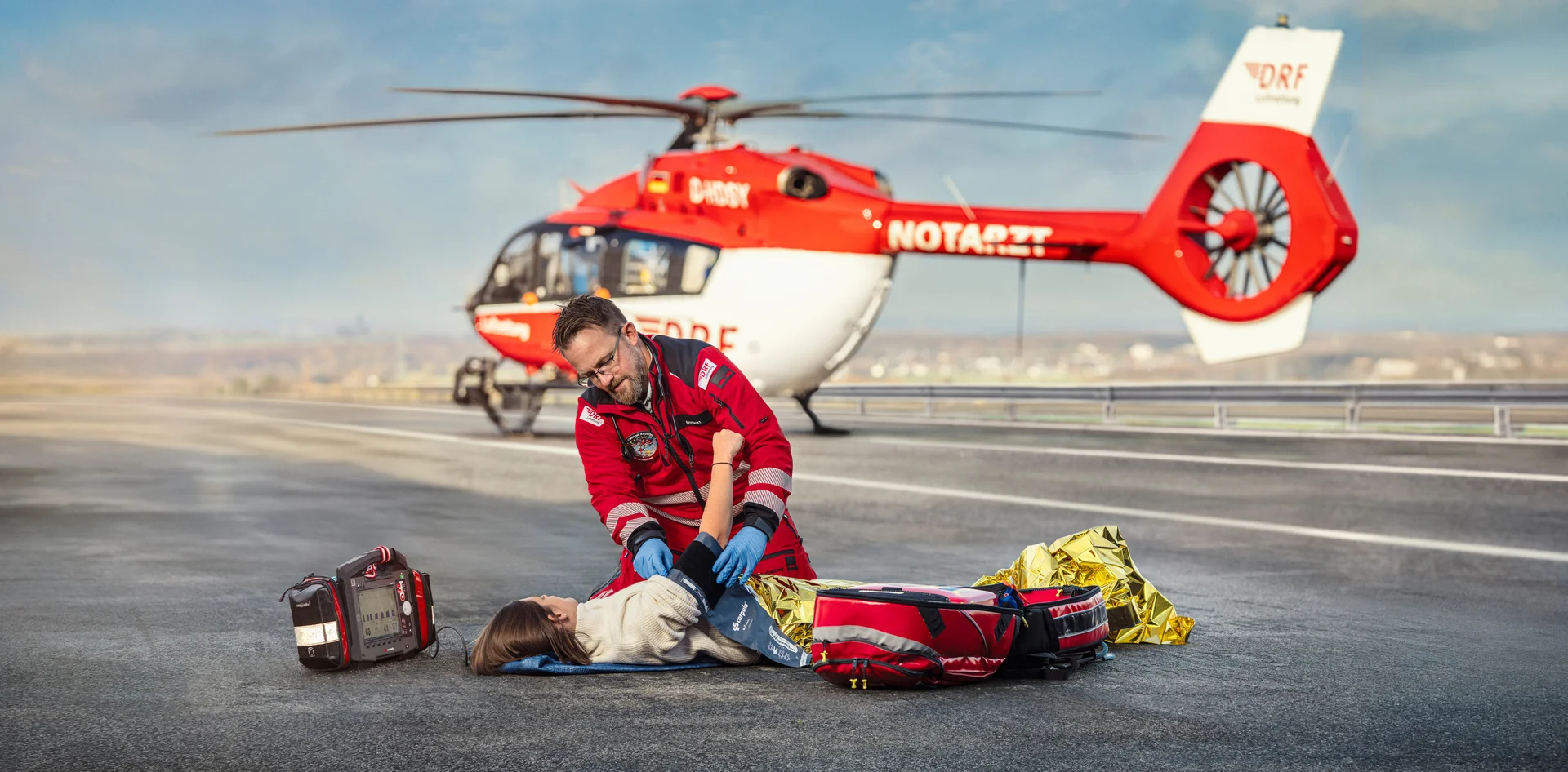 Notarzt der DRF Luftrettung versorgt eine Patientin, im Hintergrund ein rot-weißer Rettungshubschrauber.